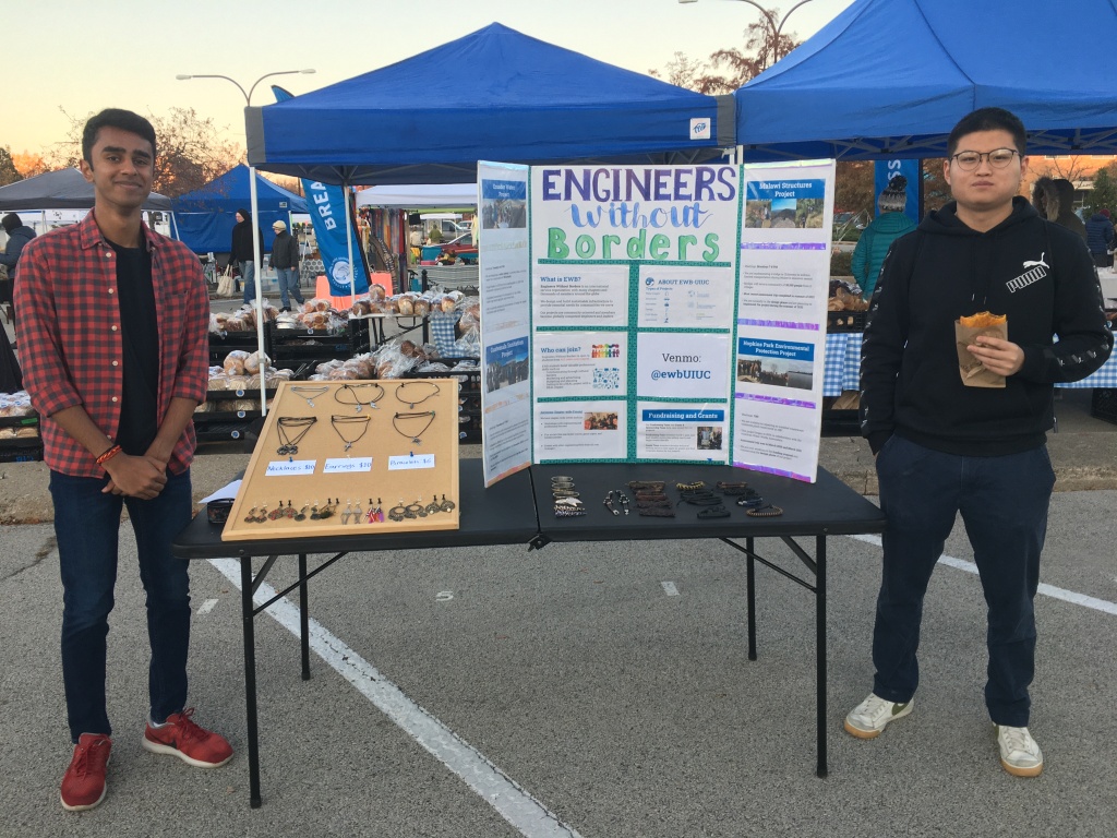 Two students standing next to a booth with necklaces, earrings and a board that reads Engineers Without Borders
